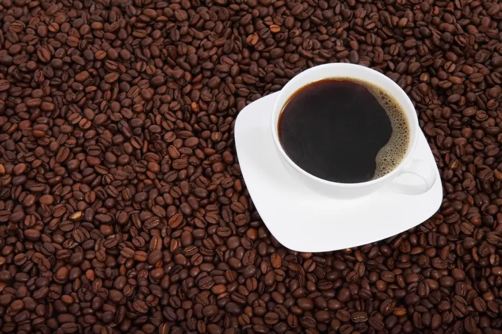 A white cup filled with black coffee placed on a surface covered with roasted coffee beans. Coffee contains caffeine, which is toxic to dogs and can cause severe health issues if consumed.